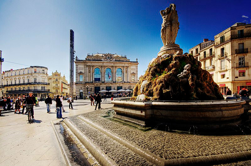 Place de la Comédie à Montpellier avec la fontaine des Trois Grâces au premier plan et l'Opéra en arrière-plan sous un ciel bleu.