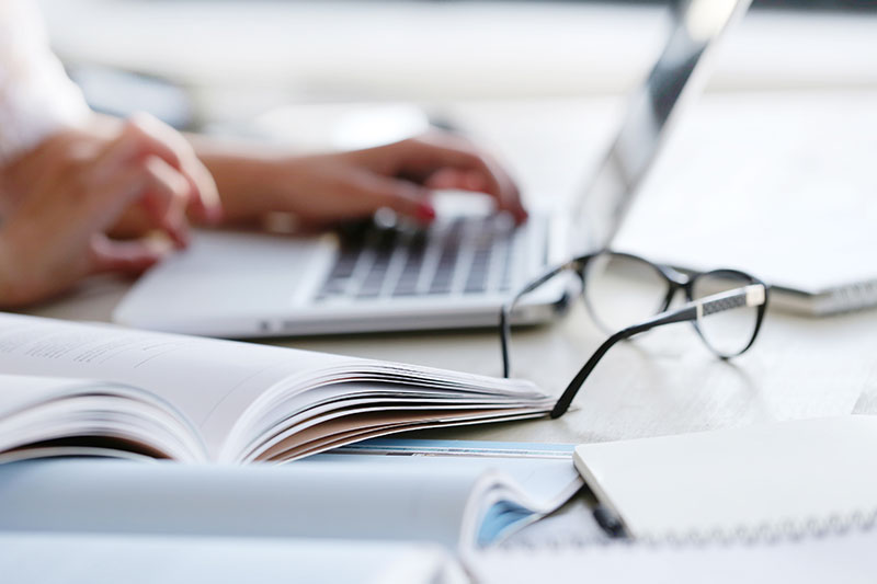 Personne utilisant un ordinateur portable avec des livres ouverts et des lunettes sur une table.
