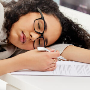 Jeune femme endormie à son bureau avec des papiers et un stylo à la main, montrant les effets du manque de sommeil.